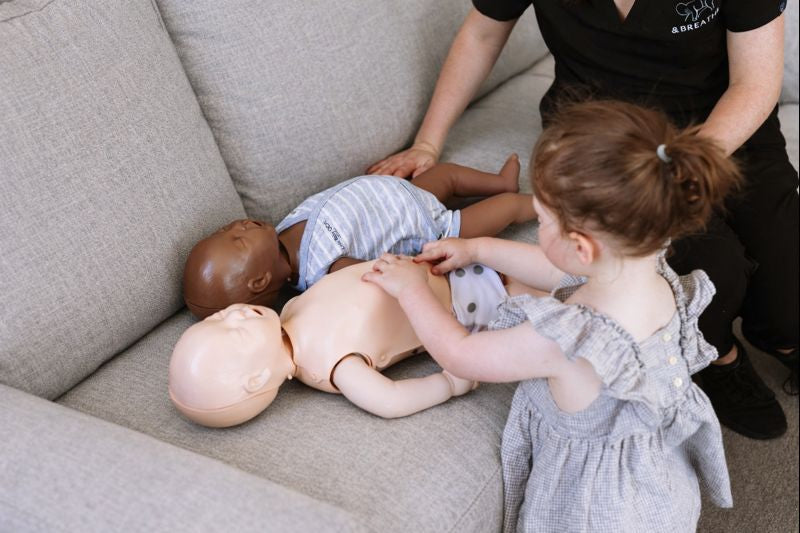 Child interacting with a mannequin on a couch, supervised by an adult at a first aid course